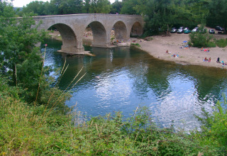 Ponte in pietra sopra il fiume a Slow Village Provence Occitane, bagnanti e auto parcheggiate sulla spiaggia.