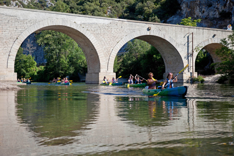 People kayaking under an old stone bridge at Slow Village Provence Occitane holiday park in Occitanie, France.