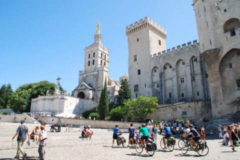 Turistas y ciclistas exploran la plaza de un castillo y iglesia histórica en Provenza, Francia, bajo el sol.