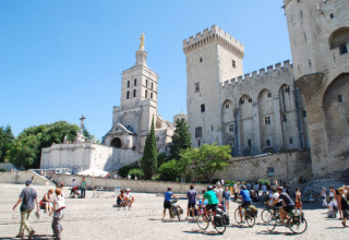 Turister og cyklister på pladsen foran et historisk slot og kirke i Provence, Frankrig, på en solrig dag.
