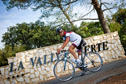 Ciclista con ropa deportiva pasando en bicicleta frente al letrero 'La Vallée Verte' en Slow Village Provence Occitane, Francia.