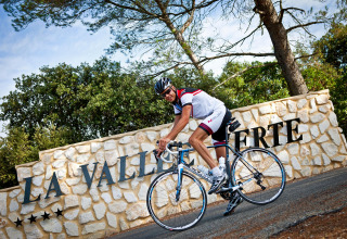 Cyklist i cykeltøj foran en stenmur med skiltet 'La Vallée Verte' i Slow Village Provence Occitane, Frankrig.