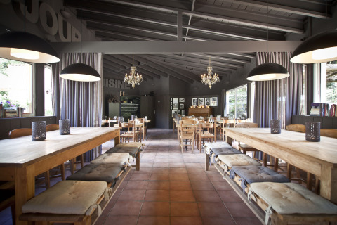 Restaurante rústico en Slow Village Provence Occitane con mesas de madera, cojines, lámparas y grandes ventanales.