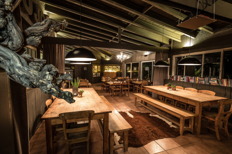 Cozy dining area with wooden tables, benches, large pendant lights, and books at Slow Village Provence Occitane, France.