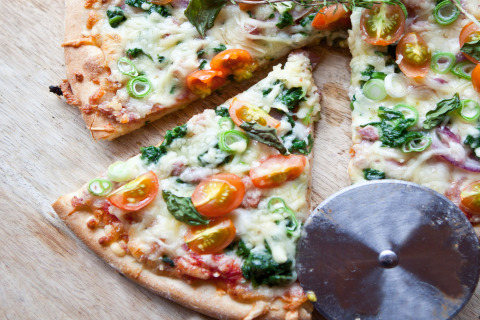 Pizza recién hecha con tomates, espinacas y cebollines, cortada en rebanadas sobre una superficie de madera.