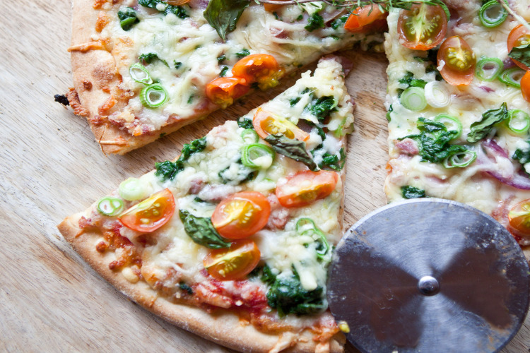 Freshly made pizza topped with tomatoes, spinach, and scallions, sliced and served on a wooden surface.