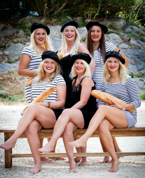 Zes dames met baretten en gestreepte jurkjes, met stokbroden, zitten blootsvoets buiten op een bank.