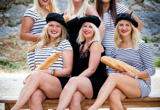 Six women in striped dresses and berets, holding baguettes, sitting barefoot on a bench outdoors.