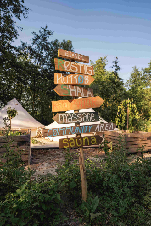 Señal artesanal de madera en RØSTIG, parque vacacional en Utrecht, Países Bajos, rodeada de vegetación.
