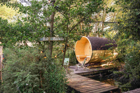 Sauna de madera tipo barril y cartel de 'Wild Wellness' en un entorno verde en RØSTIG, Utrecht, Países Bajos.