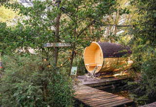Houten barrelsauna en 'Wild Wellness'-bord in het groen bij vakantiepark RØSTIG, Utrecht, Nederland.
