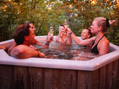Vrienden klinken met champagne in een buitenbubbelbad bij vakantiepark RØSTIG in Utrecht, omringd door bomen.