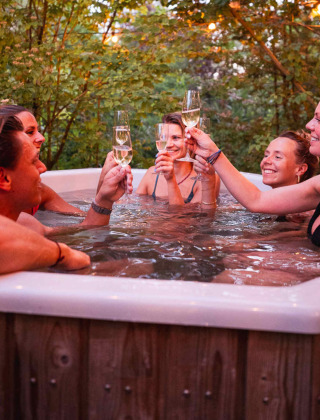 Friends toasting with champagne in an outdoor hot tub surrounded by trees at RØSTIG holiday park, Utrecht.