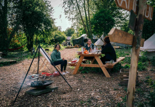 Three women enjoy a picnic at a wooden table in RØSTIG holiday park, Utrecht, Netherlands, amidst greenery.