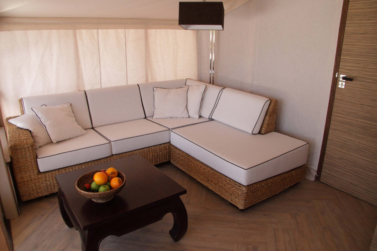 Interior de tienda safari RoyalLodge: sofá esquinero de ratán, cojines blancos y mesa con frutero.