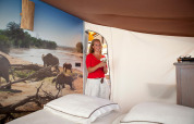Vrouw in SunLodge Safari-tent bij Slow Village Provence Occitane, Frankrijk, met olifantenmuurdecoratie.