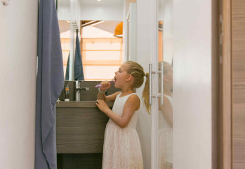 A girl in a white dress brushes her teeth in a narrow bathroom inside a SunLodge Safari tent in France.