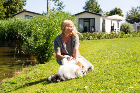 Mujer disfruta del sol con su perro en el césped junto a un lago en Recreatiepark het Esmeer, Gelderland.