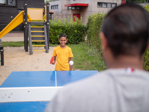 Niño jugando tenis de mesa al aire libre en Recreatiepark het Esmeer, Gelderland, Países Bajos, junto a un parque infantil.
