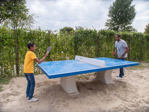 Dos personas juegan tenis de mesa al aire libre sobre arena y mesa azul en un parque vacacional de Gelderland.