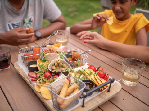 Familie genießt eine Platte mit Snacks im Freien auf einem Holztisch im Recreatiepark het Esmeer, Gelderland.