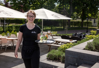 Een glimlachende serveerster brengt drankjes op het terras van Résidence Lichtenvoorde, Gelderland.