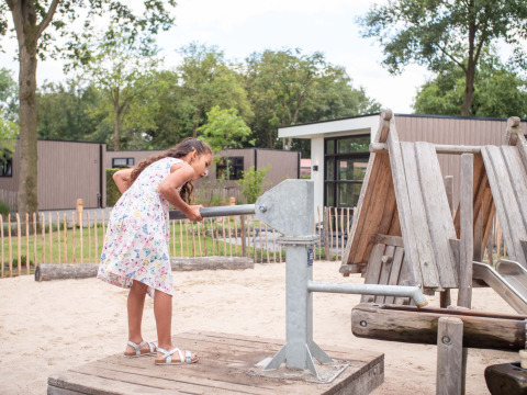 Pige i sommerkjole leger med et metalredskab på en legeplads i Résidence de Leuvert feriepark, Nord-Brabant.
