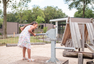 Pige i sommerkjole leger med et metalredskab på en legeplads i Résidence de Leuvert feriepark, Nord-Brabant.