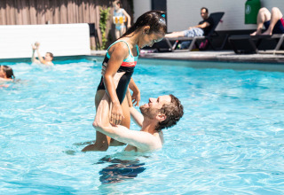 Man lifts smiling girl in swimsuit at outdoor pool in Résidence de Leuvert holiday park, North-Brabant, Netherlands.
