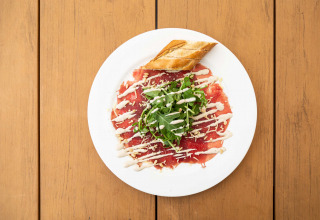 Ein Teller mit Rinder-Carpaccio, Rucola, Brot und Dressing auf Holztisch bei Résidence de Leuvert.
