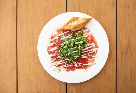 En tallerken med oksecarpaccio, rucola, brød og dressing, serveret på et træbord i Résidence de Leuvert.