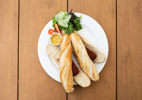 Deux baguettes avec croquettes de viande, salade verte et moutarde sur une assiette blanche en bois.