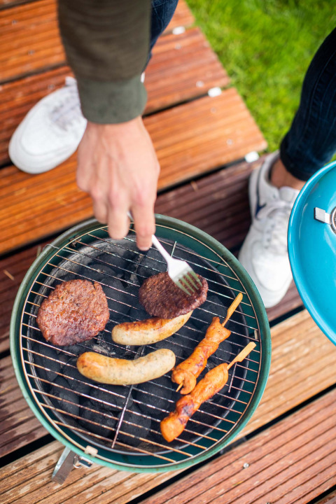 Eine Person grillt Burger, Würste und Fleischspieße auf einem kleinen Holzkohlegrill bei Landgoed de Scheleberg.