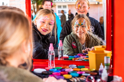 Niños con pintura facial sonríen frente a un espejo en una mesa de maquillaje colorida en Landgoed de Scheleberg.