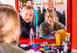 Kinder mit Gesichtsbemalung lachen vor einem Spiegel an einem bunten Schminktisch im Landgoed de Scheleberg.