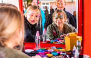 Niños con pintura facial sonríen frente a un espejo en una mesa de maquillaje colorida en Landgoed de Scheleberg.