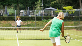 Twee mensen spelen tennis op een groene baan bij Landgoed de Scheleberg, vakantiepark in Gelderland.