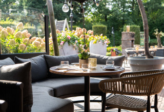 Buitenruimte met ronde tafel, grijze zetel, gevlochten stoel en bloemen in Landgoed de Scheleberg, Gelderland.