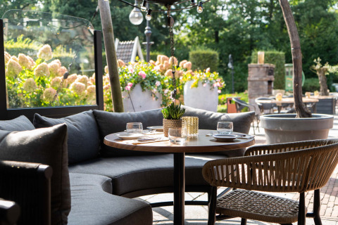 Outdoor dining area with round table, grey sofa, woven chair, and flowers at Landgoed de Scheleberg, Gelderland.