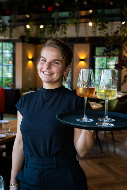 Une serveuse souriante porte deux verres de vin sur un plateau au restaurant du Landgoed de Scheleberg.