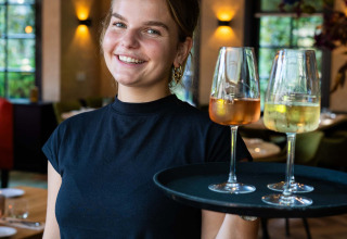 En kvindelig tjener smiler og holder en bakke med to glas vin i en restaurant på Landgoed de Scheleberg.