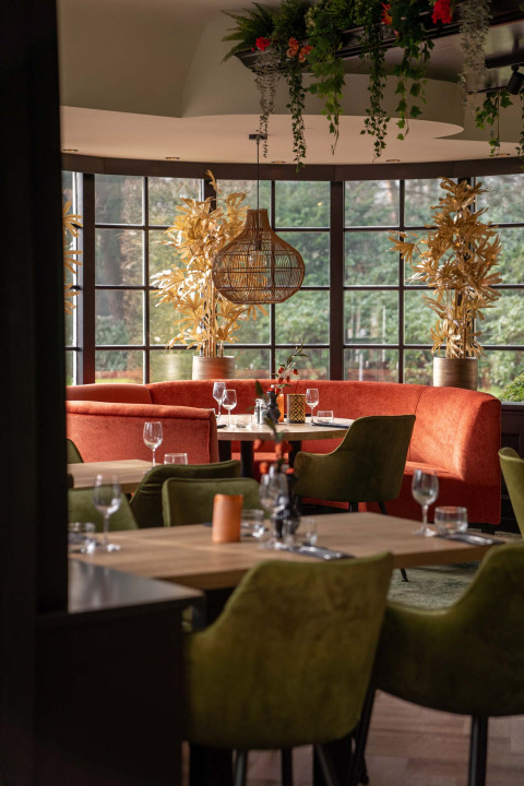Interior elegante de restaurante en Landgoed de Scheleberg, con sofás naranjas, sillas verdes y amplios ventanales.