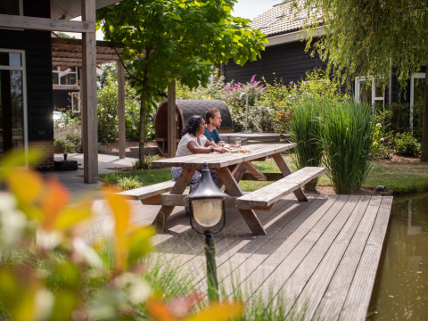Two people sit at a picnic table by the water, enjoying the sunny Résidence Lichtenvoorde in Gelderland, Netherlands.