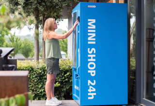 Frau steht an einem blauen MINI SHOP 24h Automaten in einem Ferienpark in Limburg, Niederlande.
