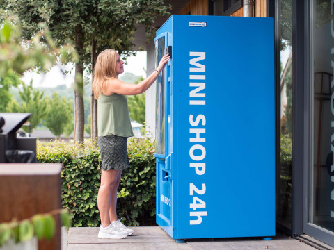 Vrouw gebruikt een blauwe MINI SHOP 24h-automaat bij een vakantiepark in Limburg, Nederland.