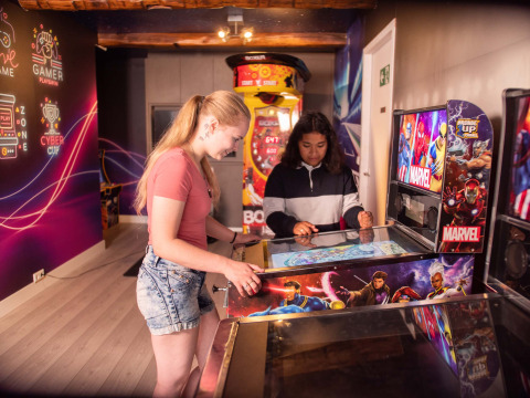 Two girls play on a Marvel pinball machine in a vibrant arcade room at Résidence Valkenburg, Netherlands.