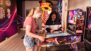 Dos chicas juegan en una máquina de pinball Marvel en una colorida sala de juegos en Résidence Valkenburg.