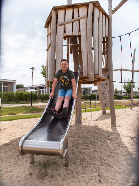 Junge rutscht auf einer Rutsche im Spielplatz vor einem Holzhaus in Résidence Valkenburg, Limburg, Niederlande.