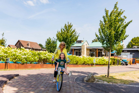 Mor og datter nyder en cykeltur på Résidence Valkenburg feriepark i Limburg, Holland, omgivet af grønne områder.