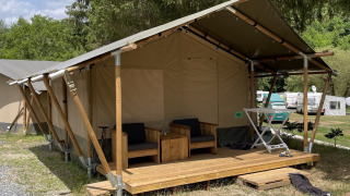 Safari tent with a wooden veranda and outdoor seating set in a grassy campground area near trees.